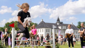 AI foto: Børn deltager i kæphestespring på Vilhelmsborg under Charity Kæpheste Cup med publikum og familieaktiviteter i baggrunden.